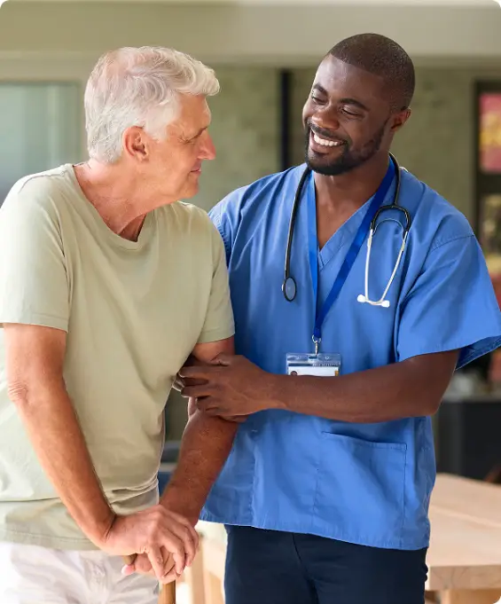 Nurse in blue scrubs smiling and supporting an elderly patient using a cane.