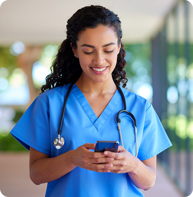 Female nurse in blue scrubs holding a smartphone and smiling, with a stethoscope around her neck in a bright hallway
