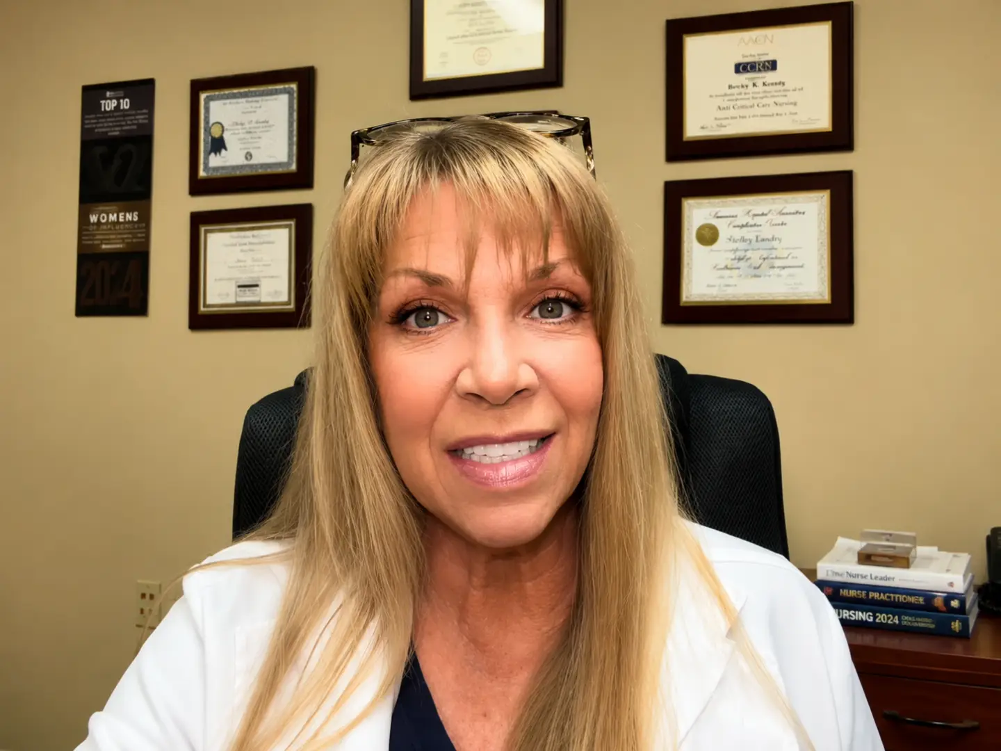 Portrait of a smiling woman with long blond hair and glasses on her head, wearing a white coat in an office with framed certificates on the wall behind her