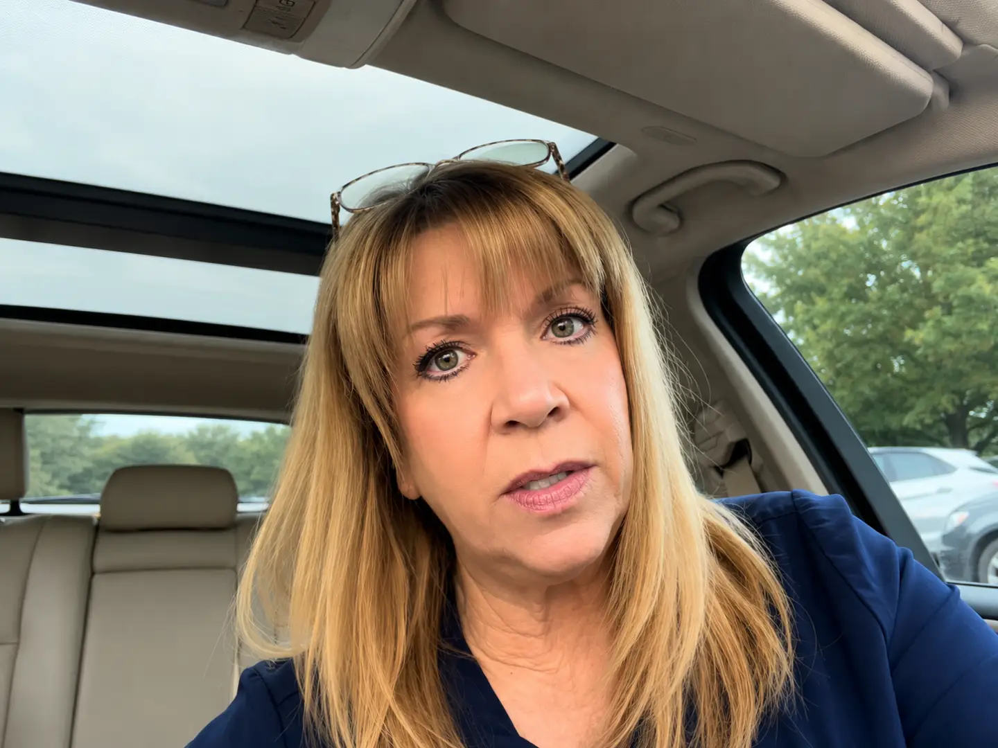 2june Woman in a car taking a selfie; sunglasses on her head, beige leather interior and sunroof visible in the background.